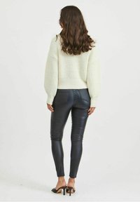 Suéter grande de punto crema con hombros caídos, combinado con leggings negros ajustados. Textura delgada y brillante con zapatos de tacón alto.