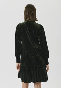 Dunkelgrünes Samtkleid mit langen Ärmeln, rundem Ausschnitt und einem gestuften Rüschensaum. Weiche Textur und geraffte Details an der Taille.