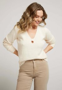 Pull en v beige clair, doux, avec des manches trois-quarts, présentant un petit accent bouton et des poignets côtelés, porté avec un pantalon beige taille haute.