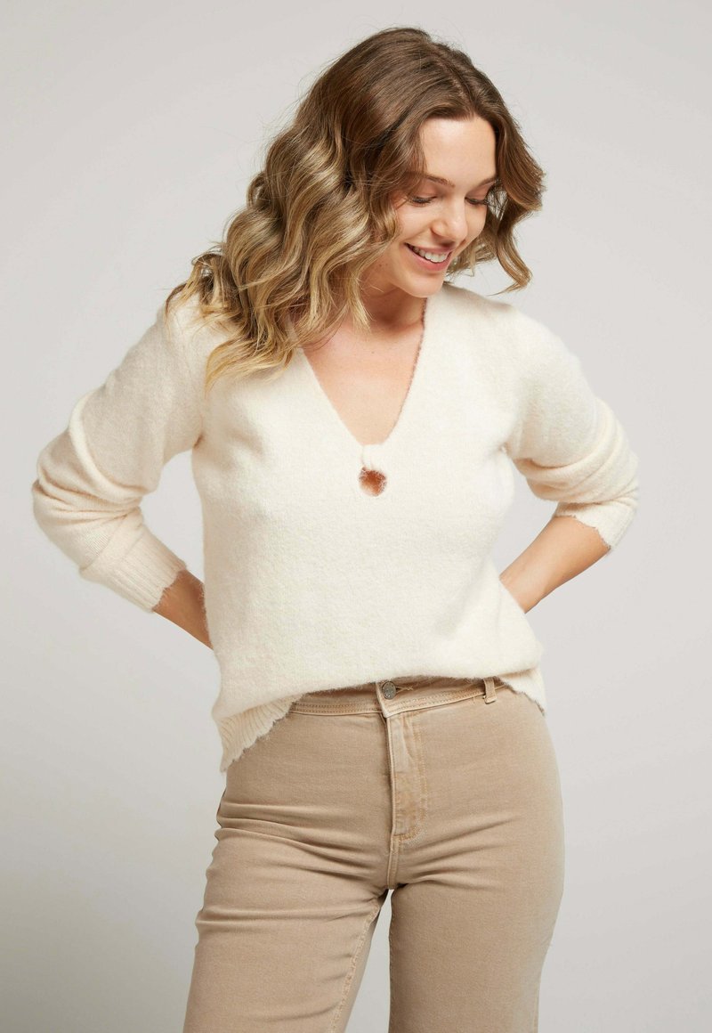 Pull en v beige clair, doux, avec des manches trois-quarts, présentant un petit accent bouton et des poignets côtelés, porté avec un pantalon beige taille haute.