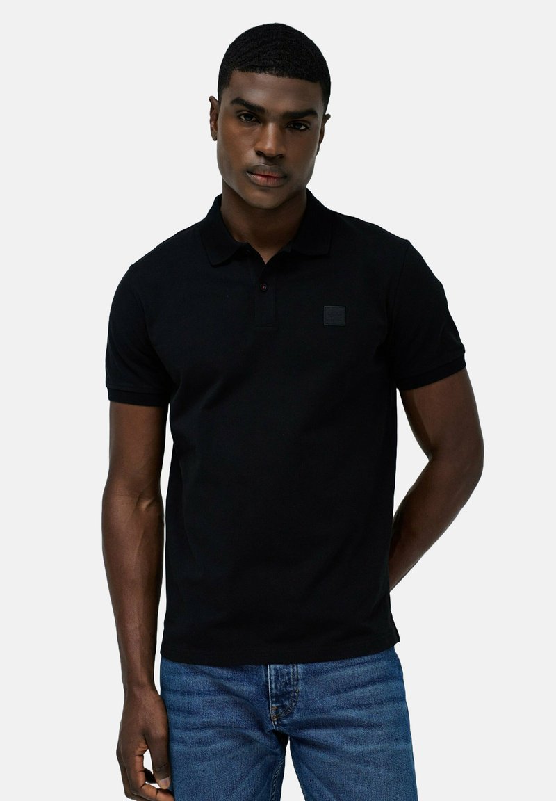 Camiseta polo negra de algodón, con cuello y mangas cortas. Pequeño parche de logo en el pecho. Combinada con jeans de mezclilla azules.