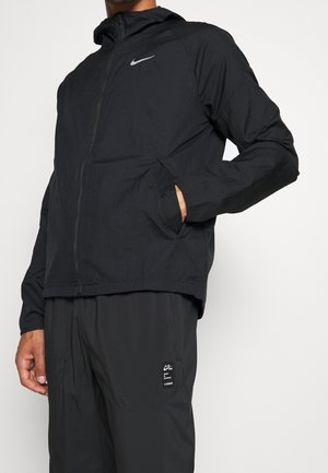 Mann trägt schwarze Nike Zip-up Jacke mit Kapuze und schwarze Nike Hose mit Etikett am Oberschenkel, steht mit einer Hand in der Jackentasche.