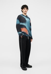 Fiatal férfi zsebre tett kézzel, kék, fekete és narancssárga mintás hosszú ujjú ingben, fekete nadrágban fehér oldalsávval és fekete cipőben áll.