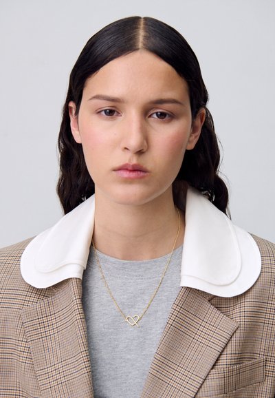 Blazer a quadri con colletto bianco, camicia grigia sotto e una delicata collana d'oro con una forma a cuore astratta.