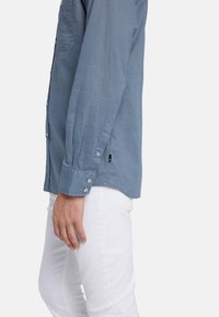 Camicia a maniche lunghe di un blu chiaro con bottoni, tasca laterale e una vestibilità comoda, abbinata a pantaloni bianchi. Il tessuto sembra morbido e testurizzato.