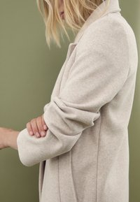 Abrigo de lana color beige claro con un diseño estructurado, mangas enrolladas y una textura suave, sobre un fondo verde.