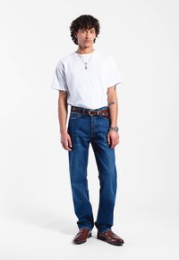 Bílé tričko s krátkým rukávem, modré džínové kalhoty, hnědý kožený pásek, hnědé boty; modelka s kudrnatými vlasy stojící před jednobarevným pozadím.