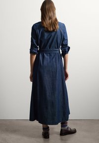 Denim-Maxikleid mit langen Ärmeln, Gürteltasche und geknöpften Manschetten. Dunkelblaue Farbe, glatte Textur. Kombiniert mit dunklen Schuhen und bunten Socken.