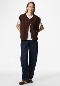 Brun, texturerad fleeceväst med knäppning framtill, bärs över en vit T-shirt. Kombinerad med löst sittande mörkblå jeans och bruna skor.