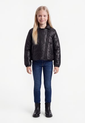Ung pige stående iført en sort skinnende jakke, blå jeans og sorte støvler, med langt blondt hår og et svagt smil.