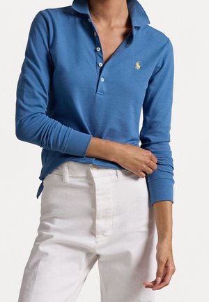 Polo à manches longues bleu avec logo jaune, associé à un pantalon blanc taille haute, bras droit du modèle croisé sur la taille.