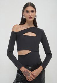 Donna con capelli neri lisci che indossa un top nero asimmetrico con ritagli e pantaloni in pelle, mostrando un tatuaggio di una rosa rossa sulla mano sinistra.