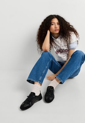 Giovane donna con capelli ricci seduta sul pavimento che indossa una t-shirt bianca con grafica, jeans blu, calzini bianchi e sneakers nere contro uno sfondo semplice.
