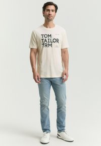 Krēmkrāsas kokvilnas T-krekls ar izteiksmīgu melnu tekstu "TOM TAILOR TRM" un akcentiem. Apvienots ar gaiši ziliem džinsiem un baltām sporta kedām.