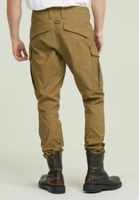 Kaki cargobroek met meerdere zakken, taps toelopende pijpen en gerimpelde boorden, gecombineerd met zwarte leren laarzen.