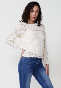 Suéter de ganchillo color crema con mangas largas, que presenta patrones geométricos y una tela texturizada. Combinado con jeans ajustados azules.