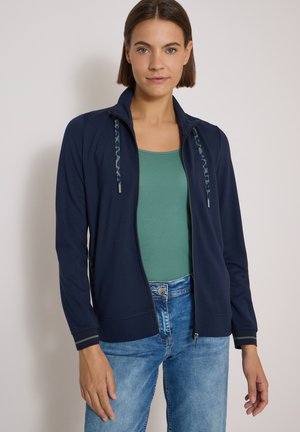 Femme aux cheveux bruns courts portant une veste zippée bleu marine sur un haut vert et un jean bleu, debout devant un mur clair et uni.