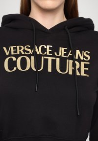 Personne portant un sweat à capuche noir avec un texte doré "Versace Jeans Couture" sur la poitrine, cordons de la capuche visibles.