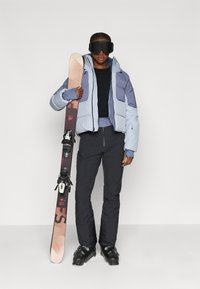 Skijaška odjeća koja se sastoji od svijetloplave i sive puffer jakne, crnog gornjeg dijela, crnih skijaških hlača i crnih skijaških čizama. Drži snowboard.