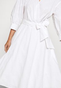 Robe blanche à manches bouffantes, avec un texte brodé "KARL LAGERFELD" et un nœud à la taille, montrée de l'avant-bras au milieu de la cuisse.