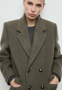 Abrigo de mezcla de lana verde oliva de doble botonadura con grandes botones negros, solapas puntiagudas y bolsillos laterales. Textura suave con un ajuste estructurado.