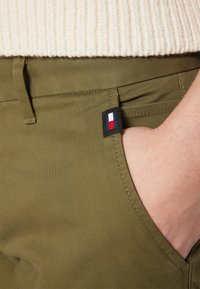 Pantaloni verde oliva con una texture liscia e una tasca laterale. Piccola etichetta in tessuto con colori blu, bianco e rosso visibile sulla tasca.