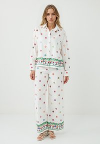 Das Model trägt ein weißes, besticktes Hemd und passende Hosen mit bunten Blumenmustern und einem grünen dekorativen Rand, posiert vor einem schlichten Hintergrund.