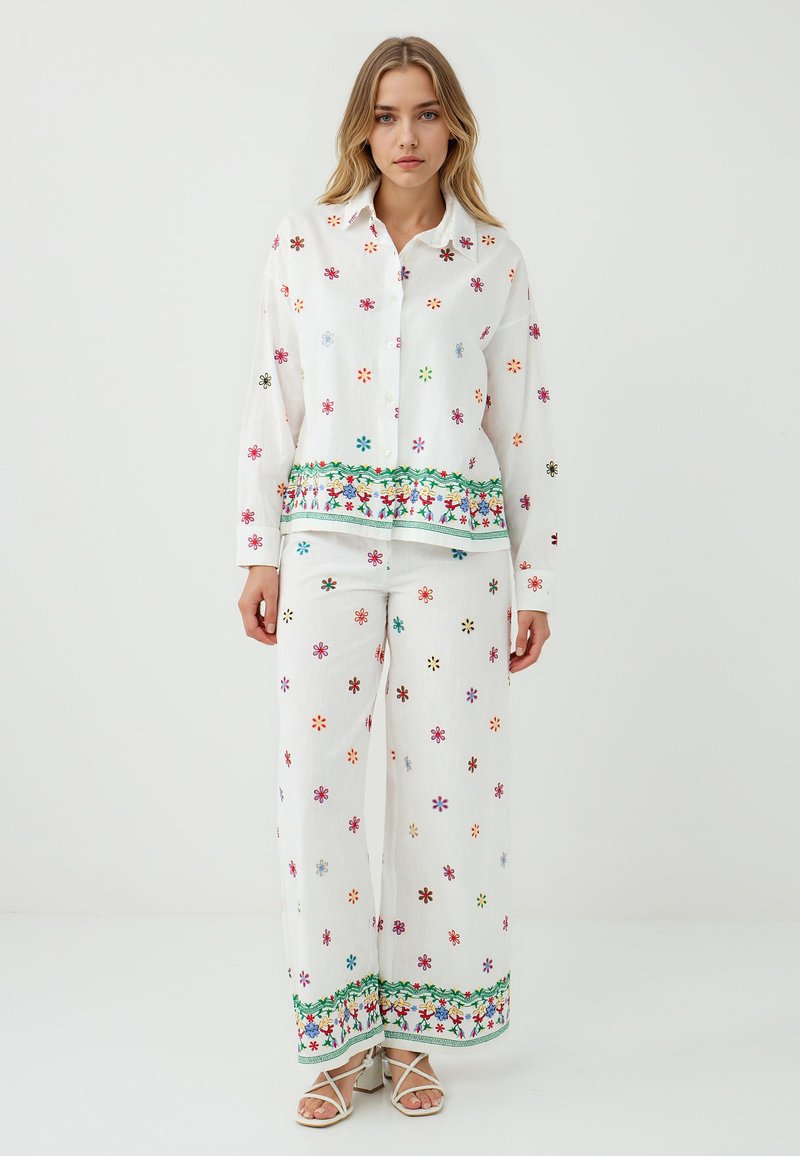 Das Model trägt ein weißes, besticktes Hemd und passende Hosen mit bunten Blumenmustern und einem grünen dekorativen Rand, posiert vor einem schlichten Hintergrund.