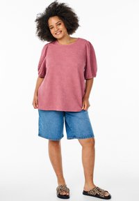 Haut rose avec un motif floral texturé et des manches bouffantes, assorti à un short en denim bleu. Sandales à motif noir et marron.