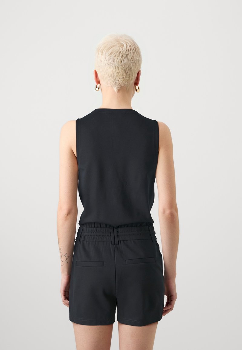 Personne aux cheveux blonds courts portant un haut noir sans manches et un short noir taille haute, debout face à un fond gris clair uni.