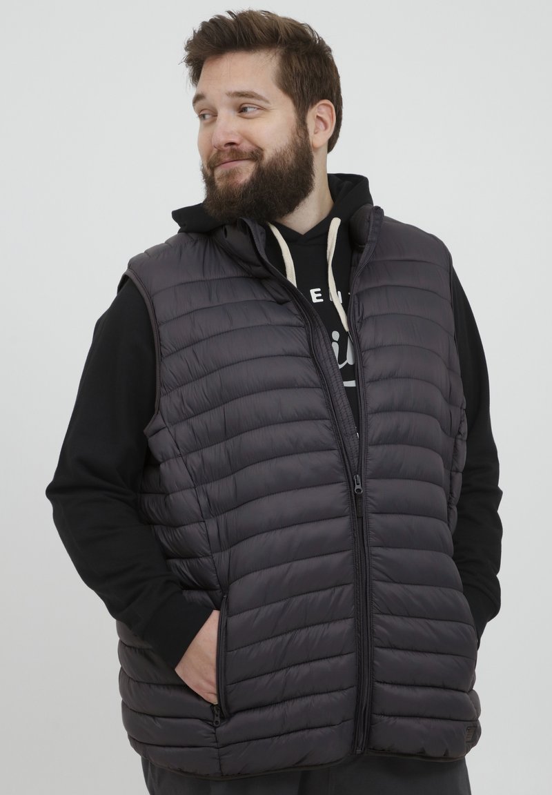 Gilet imbottito grigio scuro con zip frontale, colletto alto e tasche laterali, indossato sopra una felpa nera con cappuccio. Materiale leggero.