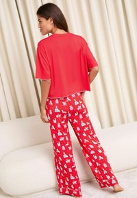 Top roșu cu mânecă scurtă, cu margine albă, purtat cu pantaloni de pijama roșii care au un imprimeu festiv repetitiv cu cadouri și animale. Material moale.