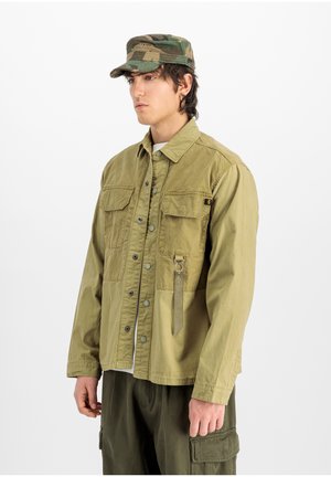 Junger Mann trägt eine khakifarbene Jacke mit Brusttaschen und einer Camouflage-Kappe, steht vor einem weißen Hintergrund.