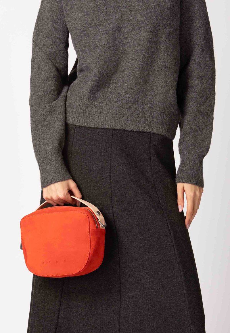 Rode suède handtas met een afgeronde rechthoekige vorm, voorzien van een lichtkleurig handvat en een ritssluiting, vastgehouden door een persoon in grijze kleding.