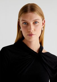 Haut noir à manches longues avec un détail de décolleté torsadé. Tissu lisse avec une silhouette ajustée. Le mannequin a de longs cheveux blonds raides et porte peu de bijoux.