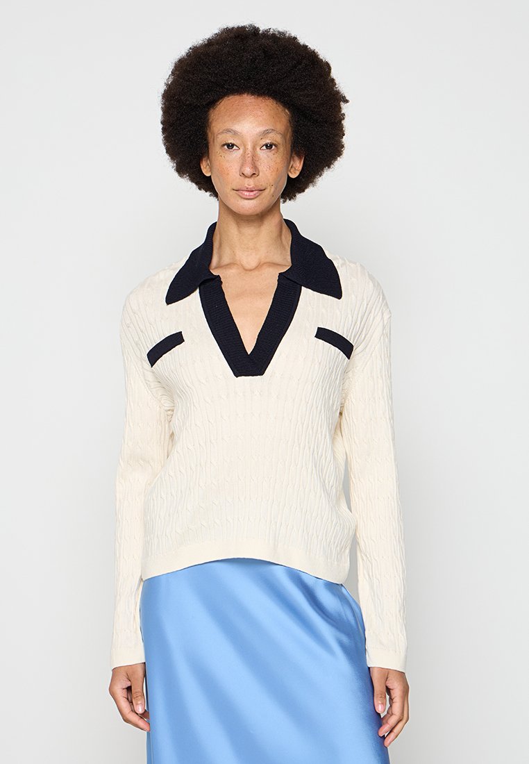 Maglione a trecce color crema con dettagli blu navy, caratterizzato da scollo a V, colletto allungato e maniche lunghe. Abbinato a una gonna di raso blu.