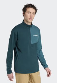 Tyrkysové tričko s dlhými rukávmi a polovičným zipsom, s ľahšou tyrkysovou vreckom a bielym logom "Terrex" na hrudi. Mäkka textúra látky.