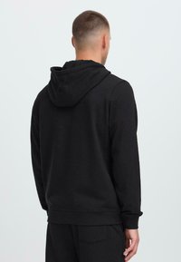 Uomo di spalle che indossa una felpa nera semplice con il cappuccio abbassato e pantaloni neri, su uno sfondo grigio chiaro.