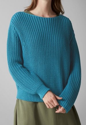 Personne portant un pull en maille épaisse bleu turquoise par-dessus une robe vert olive, ajustant la manche avec les mains.