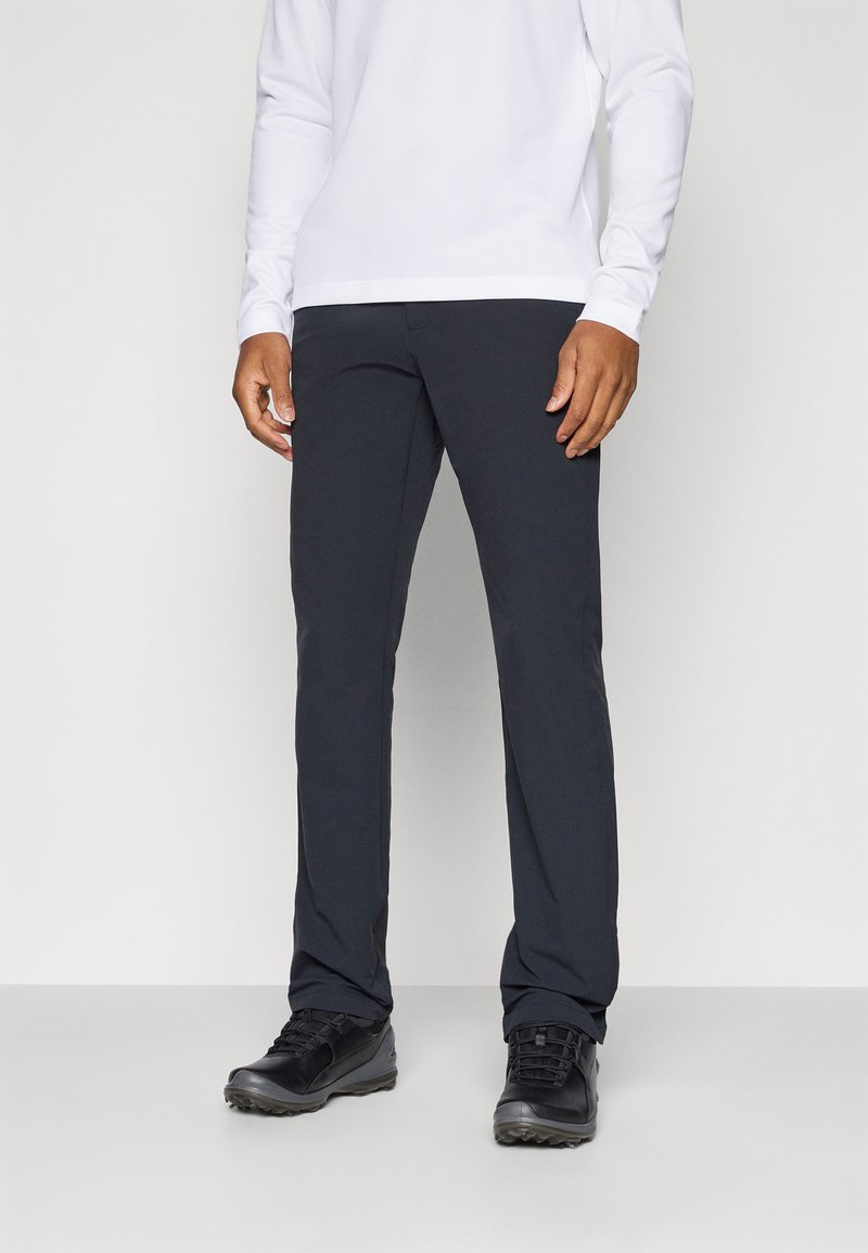 Pantalons noirs, ajustés, fabriqués dans un tissu lisse et extensible. Assortis avec un haut à manches longues blanc et des chaussures de plein air noires. Pas de motifs visibles.