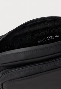 Vnitřek černého nylonového pytle s hladkou texturou, obsahující kapsu na zip a viditelnou značkovou etiketu "Marc O'Polo".