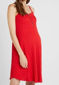 Robe rouge à côtes avec des bretelles spaghetti réglables et un devant boutonné. Tissu fluide avec un ourlet à volants. Adaptée à diverses occasions.