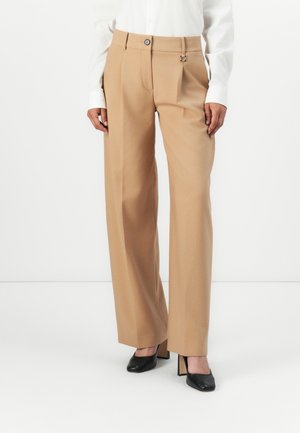 Femme portant un pantalon beige à jambes larges avec un détail bouton et boucle, associé à des chaussures à talons noires et une chemise blanche.