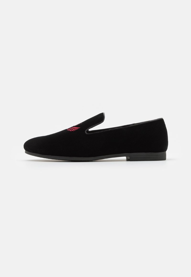 Mocassin en velours noir sans lacets avec semelle plate et petit détail d'aile rouge brodé sur le dessus, présenté de profil sur fond blanc.