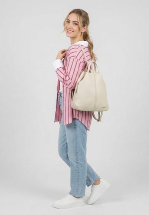 Jonge vrouw glimlachend, gekleed in een roze gestreept shirt, lichtblauwe jeans en witte sneakers, met een beige leren rugzak op haar rug.