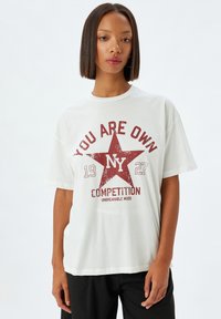 Camiseta blanca de algodón con estampado gráfico rojo que presenta una estrella, el texto "TÚ ERES PROPIO" y "COMPETENCIA". Cuello redondo, mangas cortas, corte relajado.