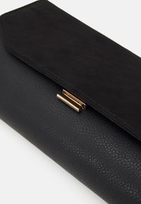 Black clutch purse featuring textured faux leather, a smooth suede flap, and a gold-tone clasp detail. Compact and rectangular shape.