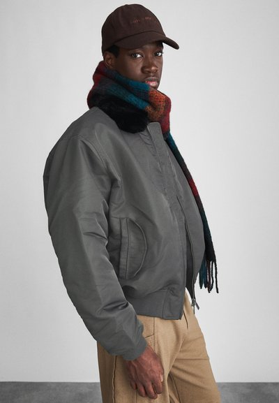 Veste bomber grise avec fermeture éclair et deux poches latérales, superposée à une écharpe à carreaux multicolores. Le mannequin porte une casquette marron foncé.
