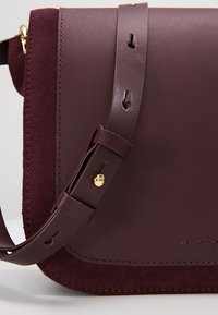 Borsa in pelle e suede bordeaux con chiusura a pattina, una tracolla sottile e regolabile, e dettagli in acciaio dorato. Texture liscia con dettagli di cucitura.