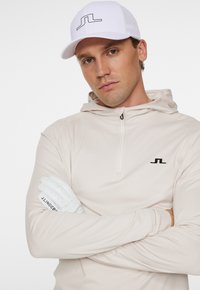Ljust beige zip-up hoodie med svart logotyp på bröstet, kombinerad med vita fingerlösa golfhandskar. Modellen bär en vit keps med en svart linje-logotyp.